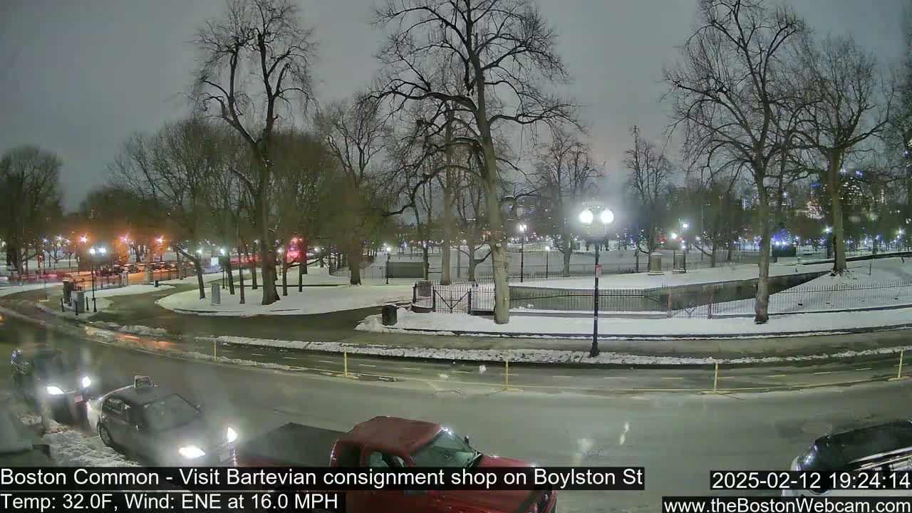 A nighttime view of a snow-dusted park with cars parked along a road, under a cloudy sky with a wind of 16 mph.