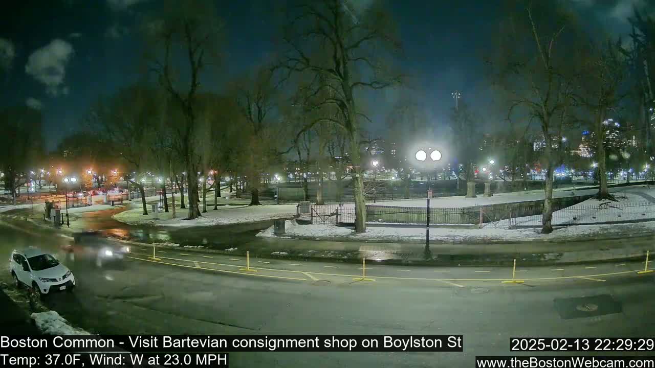 A nighttime view of a snow-covered park with a road and cars, under a partly cloudy sky, with a strong westerly wind.