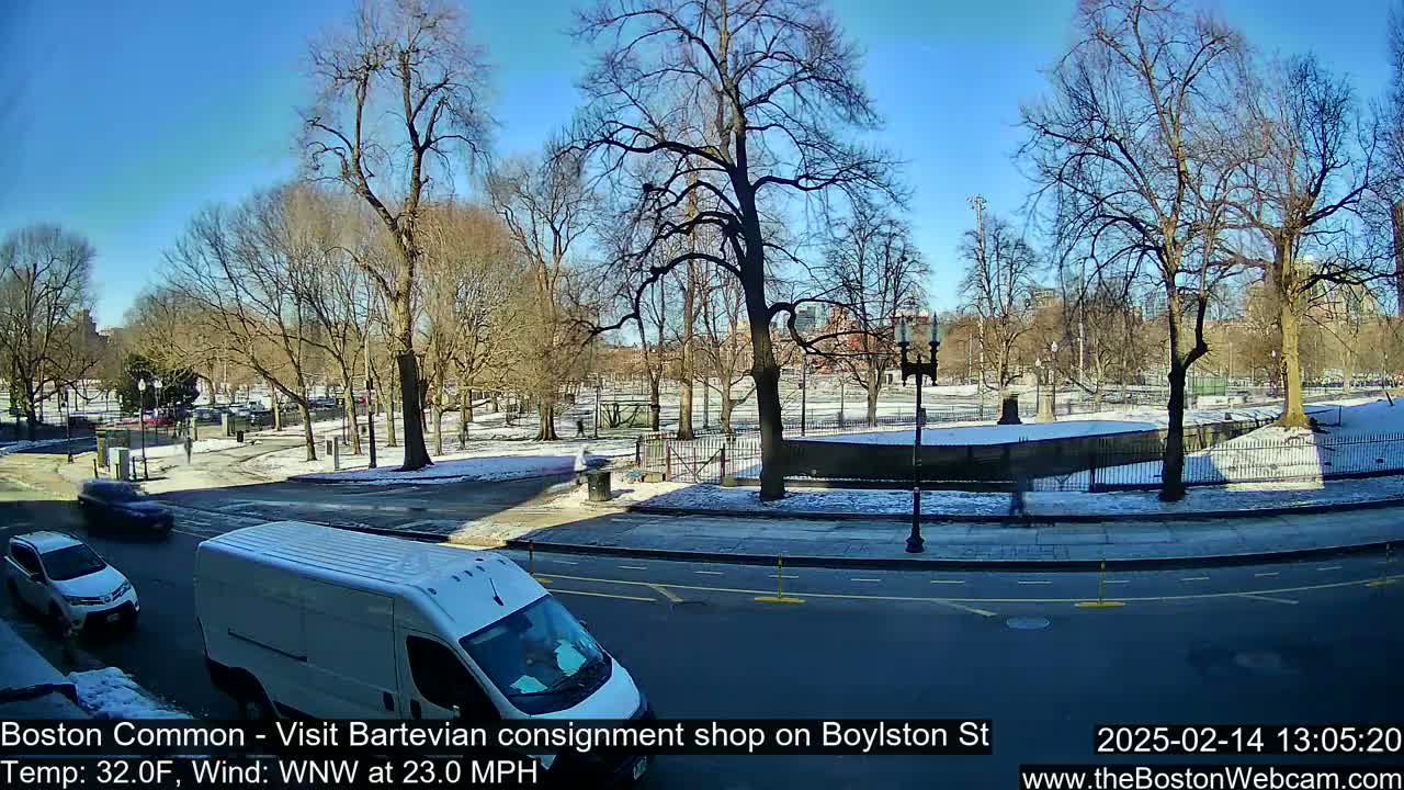A sunny, cold day with snow on the ground shows a city street with cars driving past a park enclosed by a fence with bare trees.