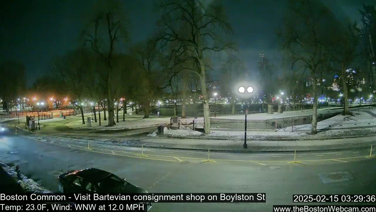 A nighttime view of a snow-dusted park with a road and cars, under a dark sky with a wind from the west-northwest at 12 miles per hour and a temperature of 23 degrees Fahrenheit.