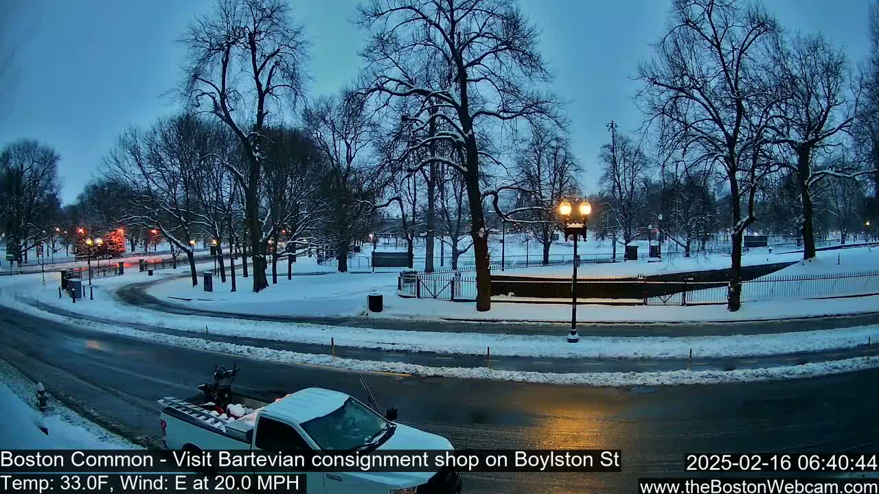 A snow-covered park with bare trees and streetlights at dusk, a partially visible white truck is parked on the road next to it, and the road is partly covered in snow.