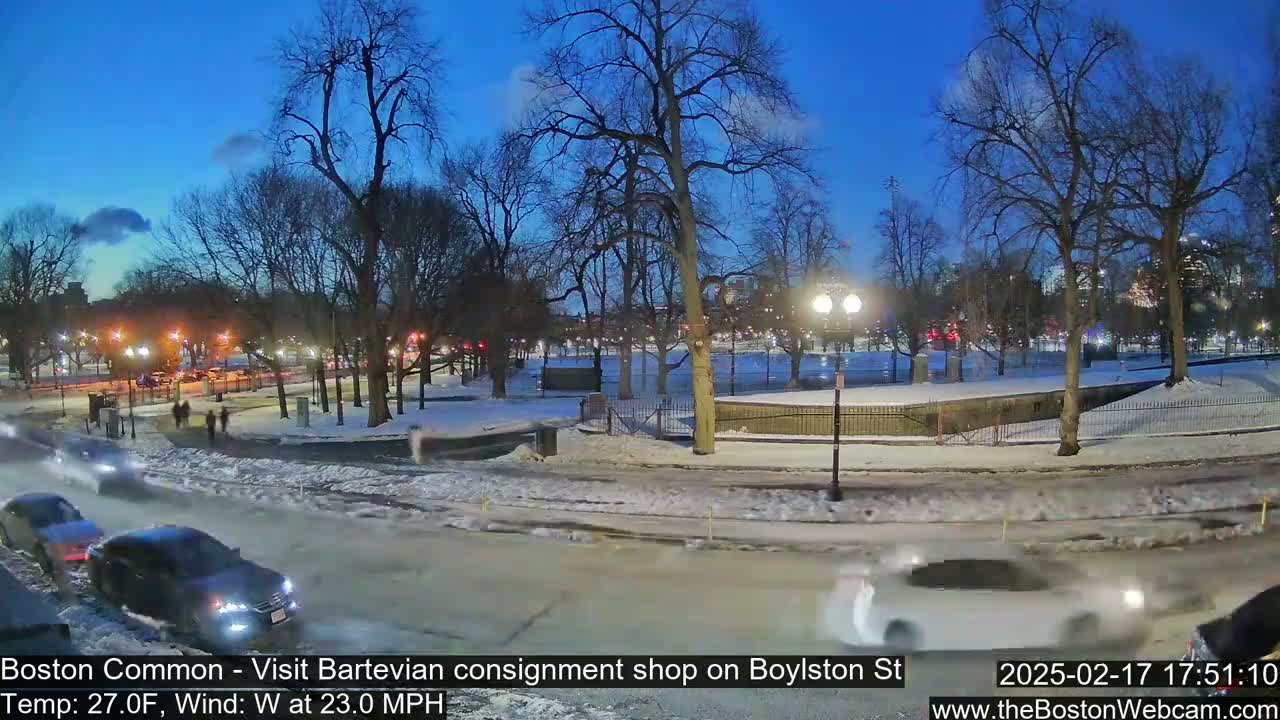 A snow-covered park at dusk with cars driving on a nearby road, under a clear, twilight sky and a 23 mph westerly wind.