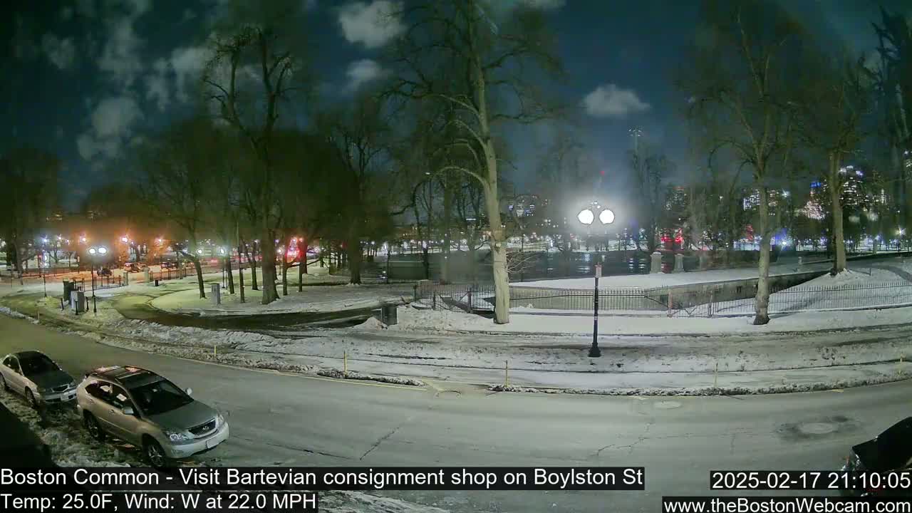 A snow-covered park at night, under a partly cloudy sky, with a few cars parked along a street nearby.