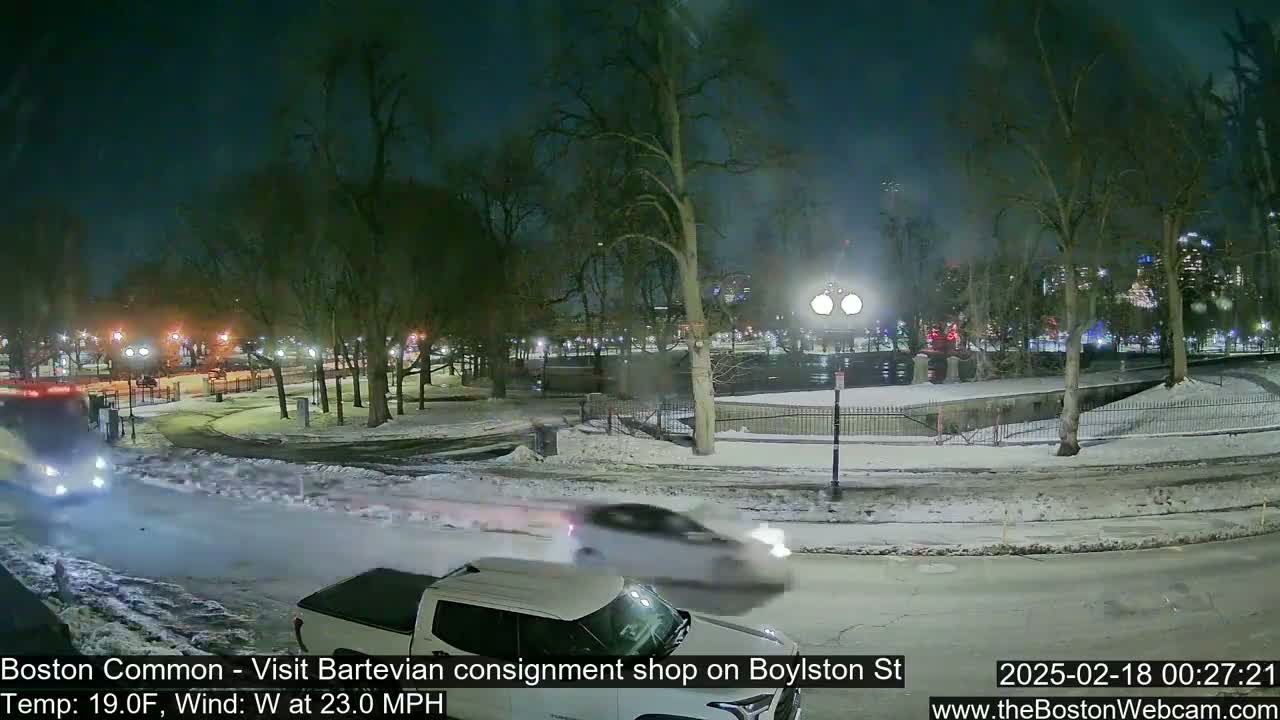 A snow-covered park at night with a few vehicles driving on a nearby road under a partly cloudy sky, with a strong west wind.