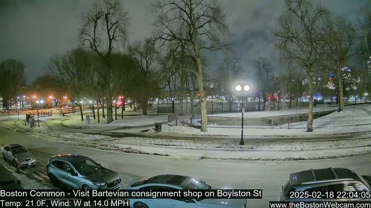 A nighttime view of a snow-covered park with several cars parked along a road, under a cloudy sky with a westerly wind.