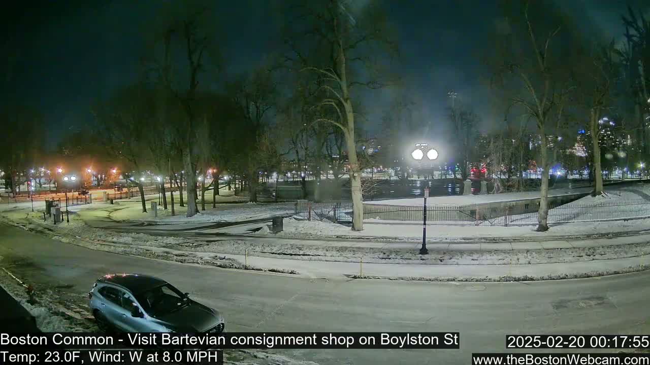 A nighttime view of a snow-covered park with a car parked on a street, under a dark sky with a westerly wind.