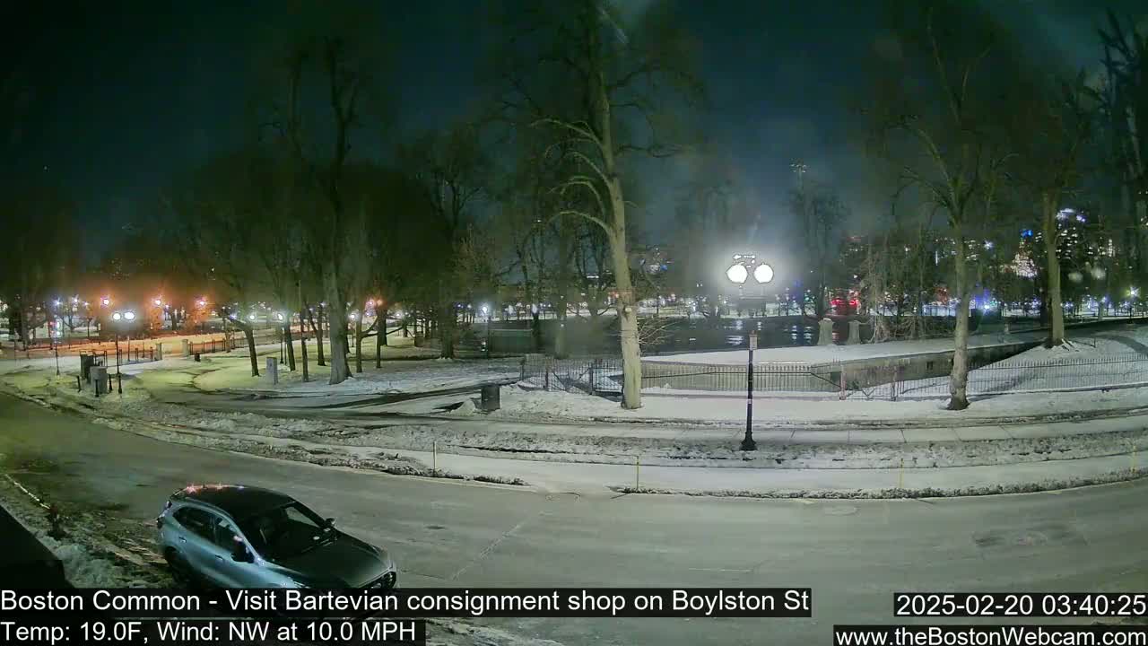 A snow-covered park at night, illuminated by streetlights, with a car parked on a nearby road, under a dark, clear sky with a northwest wind.