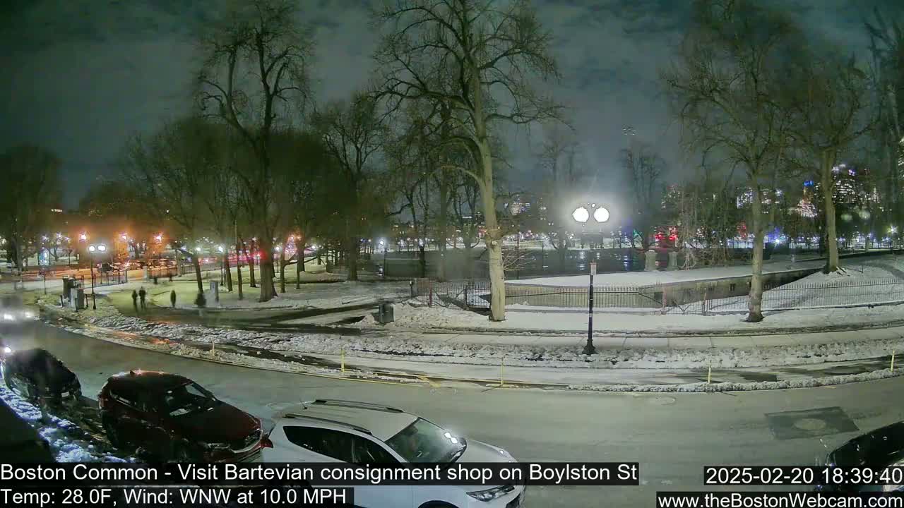 A nighttime view of a snow-covered park with cars parked along a street, under a cloudy sky.