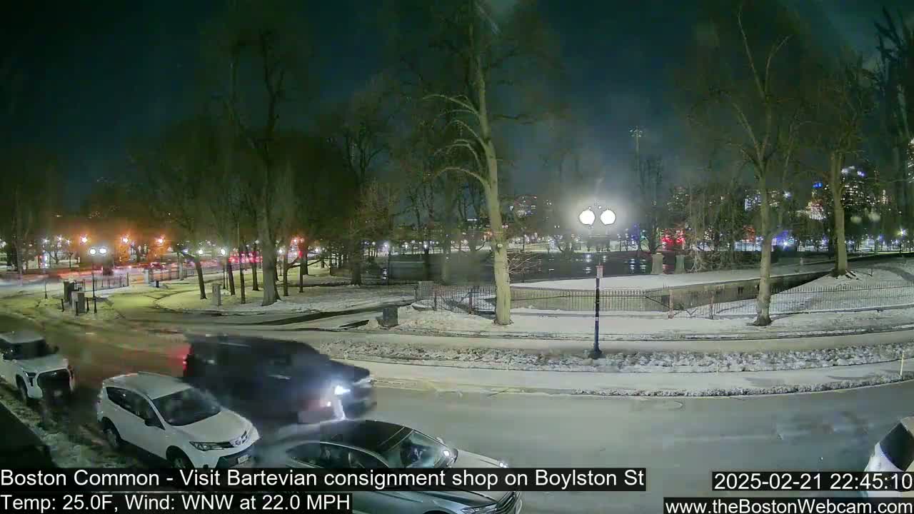 A nighttime scene of a snow-covered park with cars driving on a nearby road, under a dark sky with a wind blowing from the west-northwest at 22 mph.