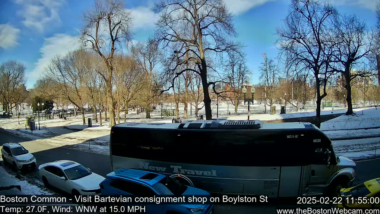 A partly sunny day with snow on the ground shows a city park with bare trees and several cars and a bus on a nearby street.
