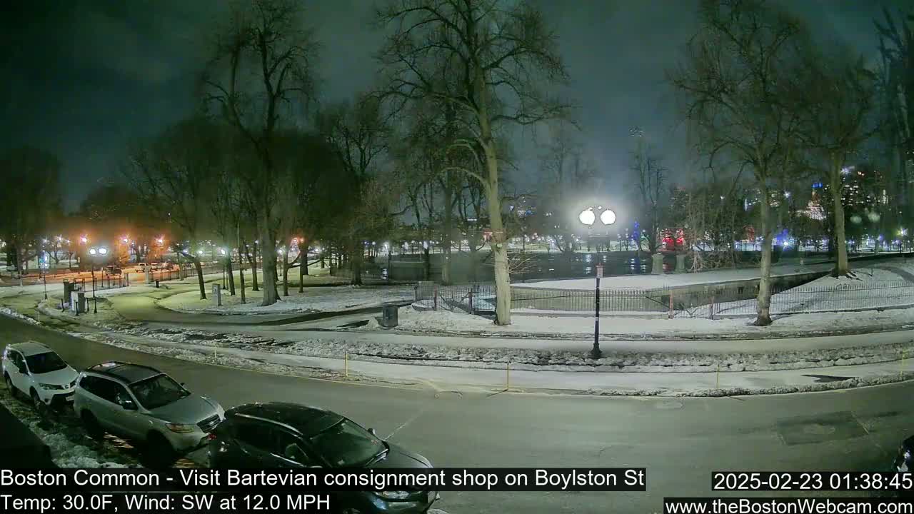 A snow-covered park at night, with cars parked along a road beside it, under a dark, somewhat cloudy sky.
