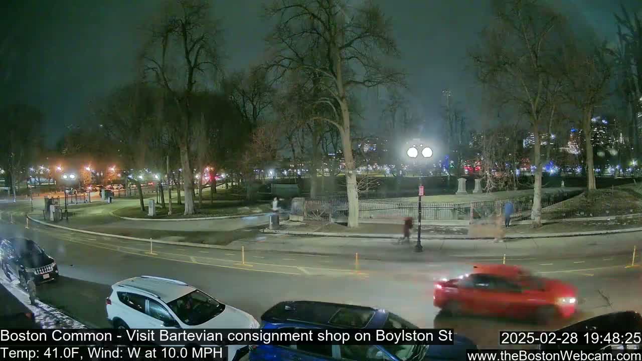 A nighttime scene shows a park with bare trees surrounding a paved roadway where several cars are parked and driving, under a cloudy sky.