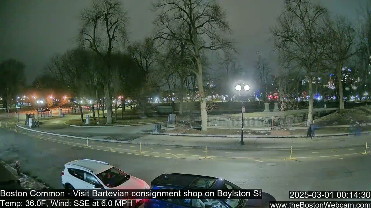 A nighttime view of a park, with a few people walking and cars parked along a roadway, under a cloudy sky with a wind from the southeast at 6 mph.