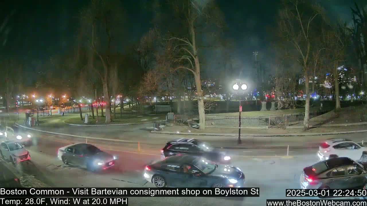 Several cars drive along a road at night past a park with bare trees, under a windy, 28-degree Fahrenheit sky.