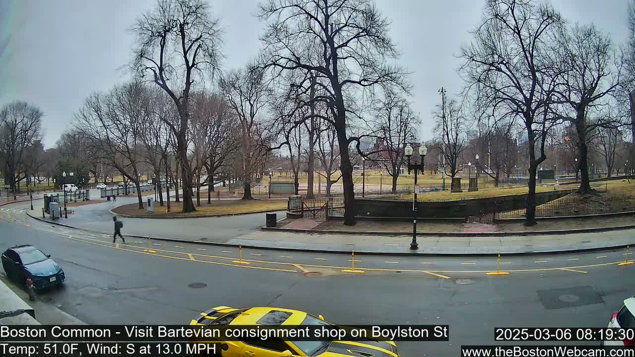 A partly cloudy, cool day shows a person walking near a road bordering a park with bare trees and a yellow car parked near the road.