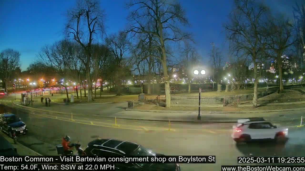 A nighttime view of a park with bare trees, cars on a road, and people walking, under a clear, dark blue sky with a strong south-southwesterly wind.