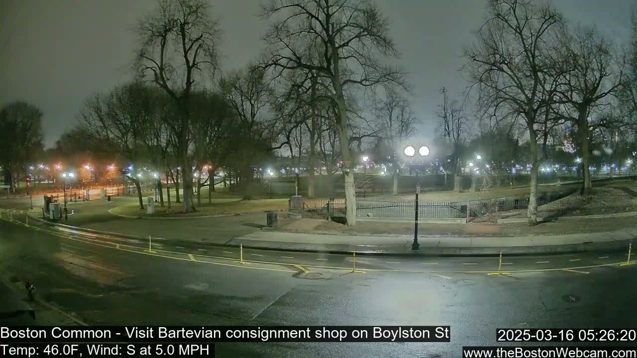 A nighttime view of a park with bare trees, lit pathways, and a road, under a cloudy sky with a south wind.