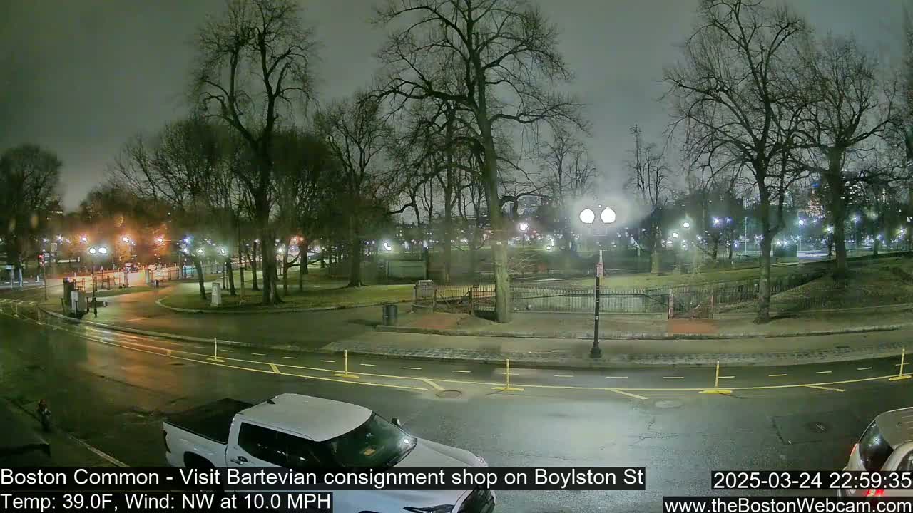 A nighttime view of a park with bare trees, illuminated by streetlights, under a cloudy sky, with a wet road and a white truck partially visible in the foreground.