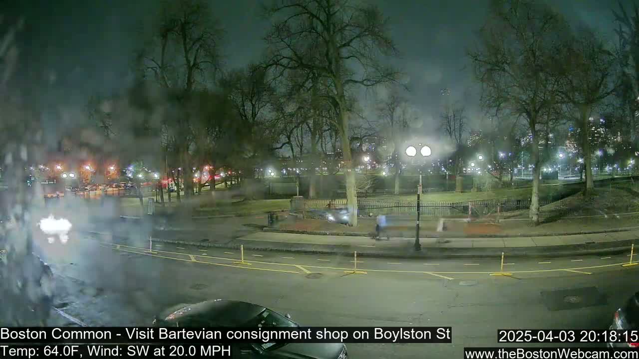 A nighttime view of a park and street, with bare trees, a few pedestrians, and cars, under slightly hazy conditions and a southwest wind.