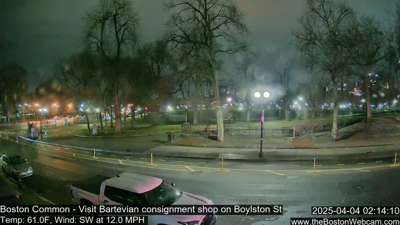 A nighttime view of a park with a few cars on a wet street, under a dark, overcast sky with a southwest wind.