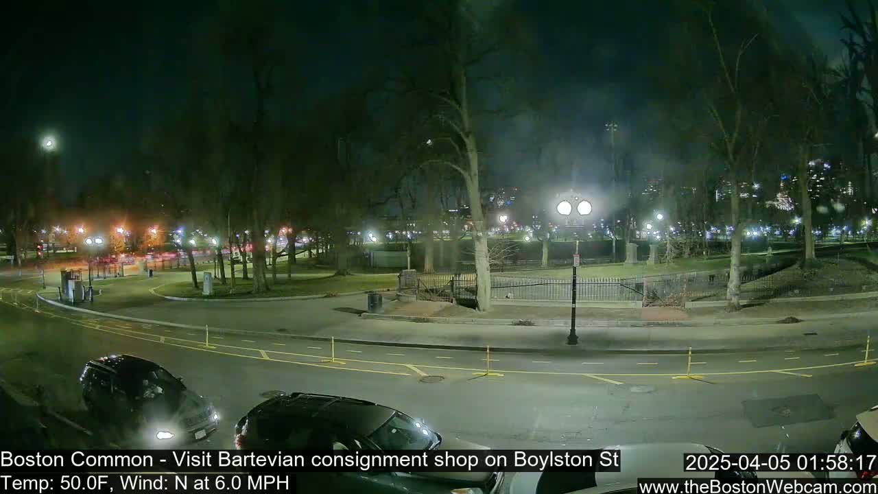 A nighttime view of a park and street, with a few cars parked and driving, under a clear sky with a 50°F temperature and a 6.0 mph north wind.