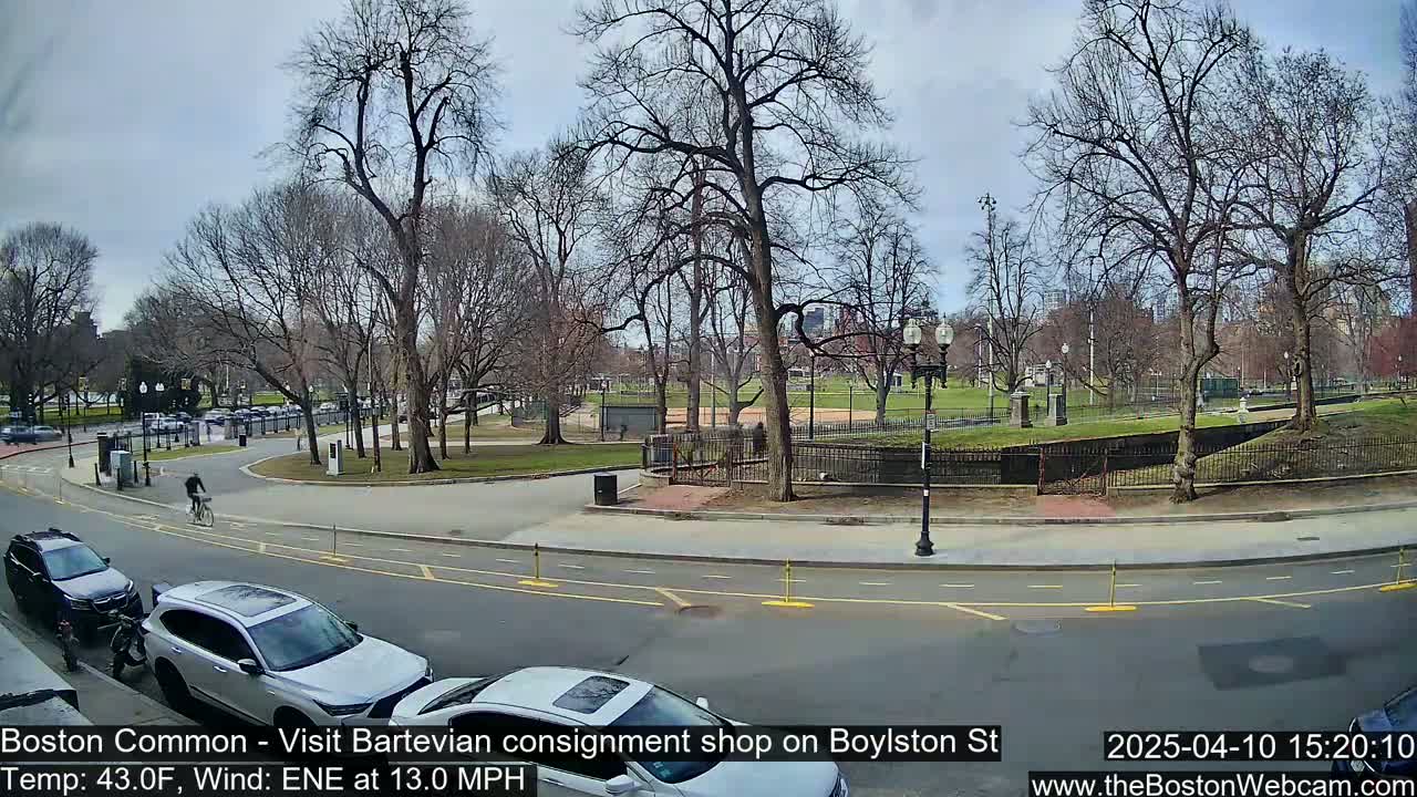 A partly cloudy day shows a cyclist riding past cars parked near a park with bare trees and a grassy area.