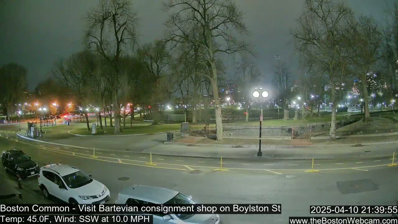 A nighttime view of a park with a few cars parked on the street, under a cloudy sky with a 10 mph wind.