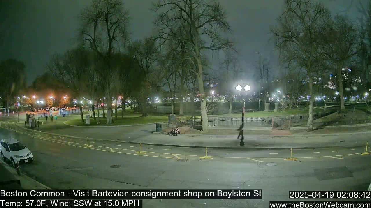 A nighttime view of a park and street, with a few people visible, under a cloudy sky and a 15 mph wind.