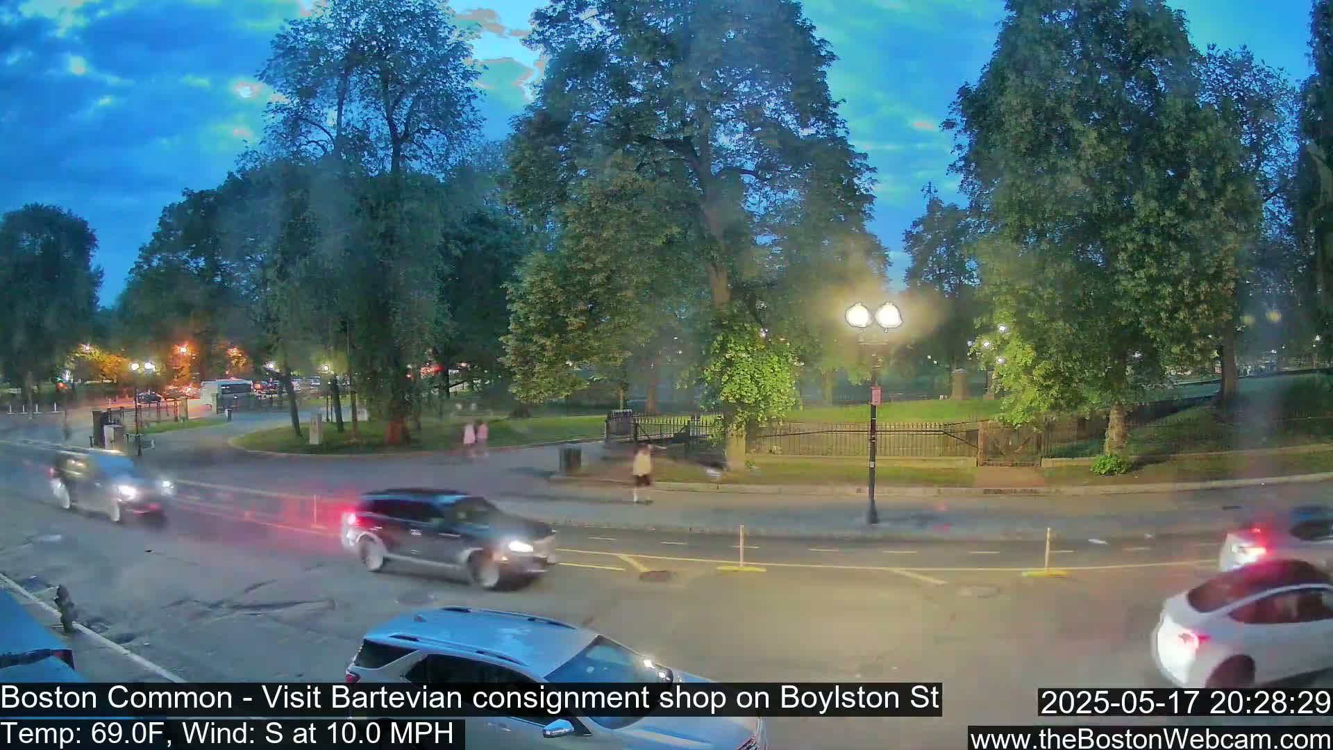 Cars drive along a street at dusk, past a park with lush green trees under a mostly clear, twilight sky with a light south wind.