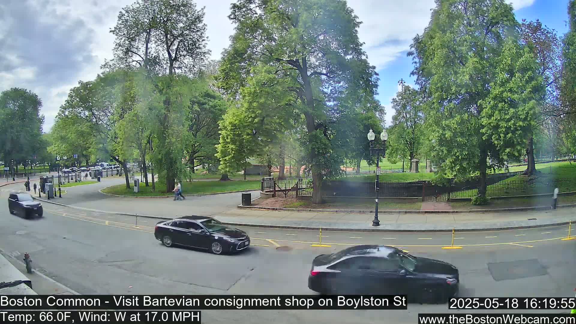 A partly sunny street scene shows cars driving past a park with lush green trees and grass, under a partly cloudy sky.