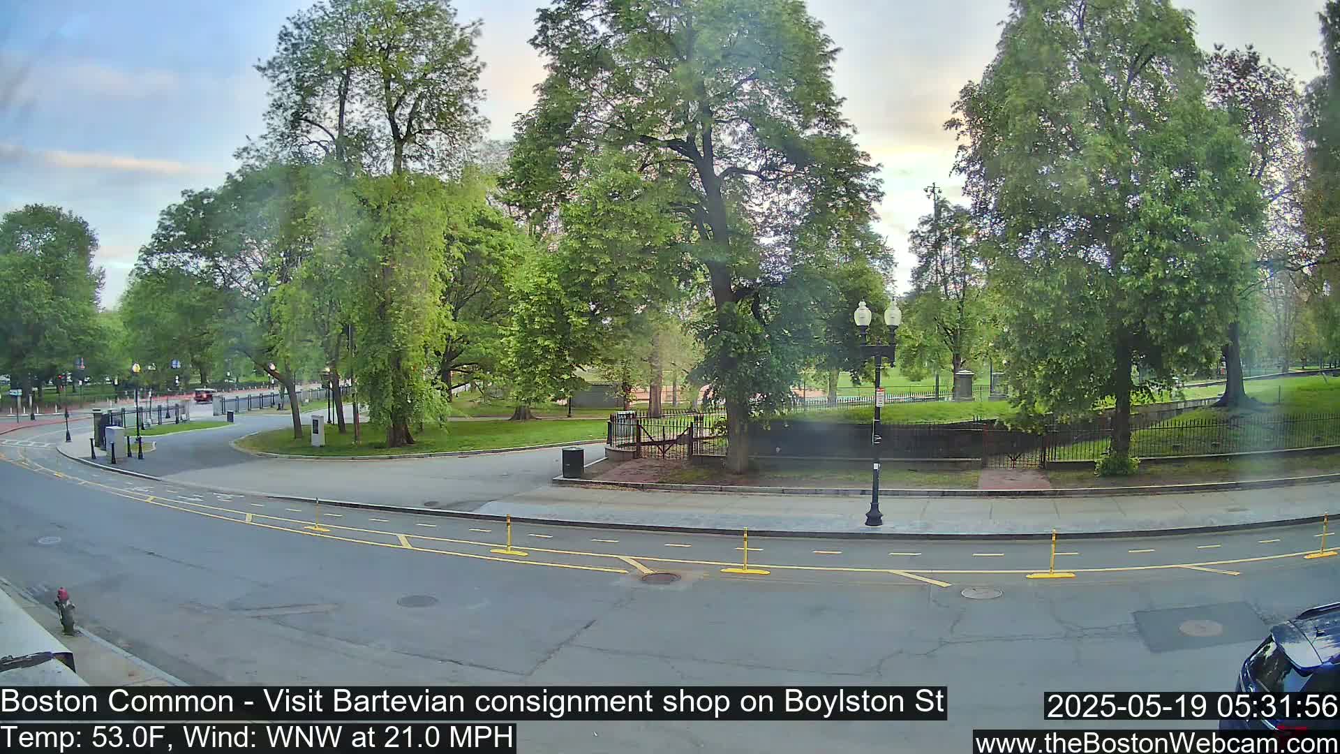 A street scene shows a roadway curving past a park with lush green trees under a partly cloudy sky, with a gentle breeze.
