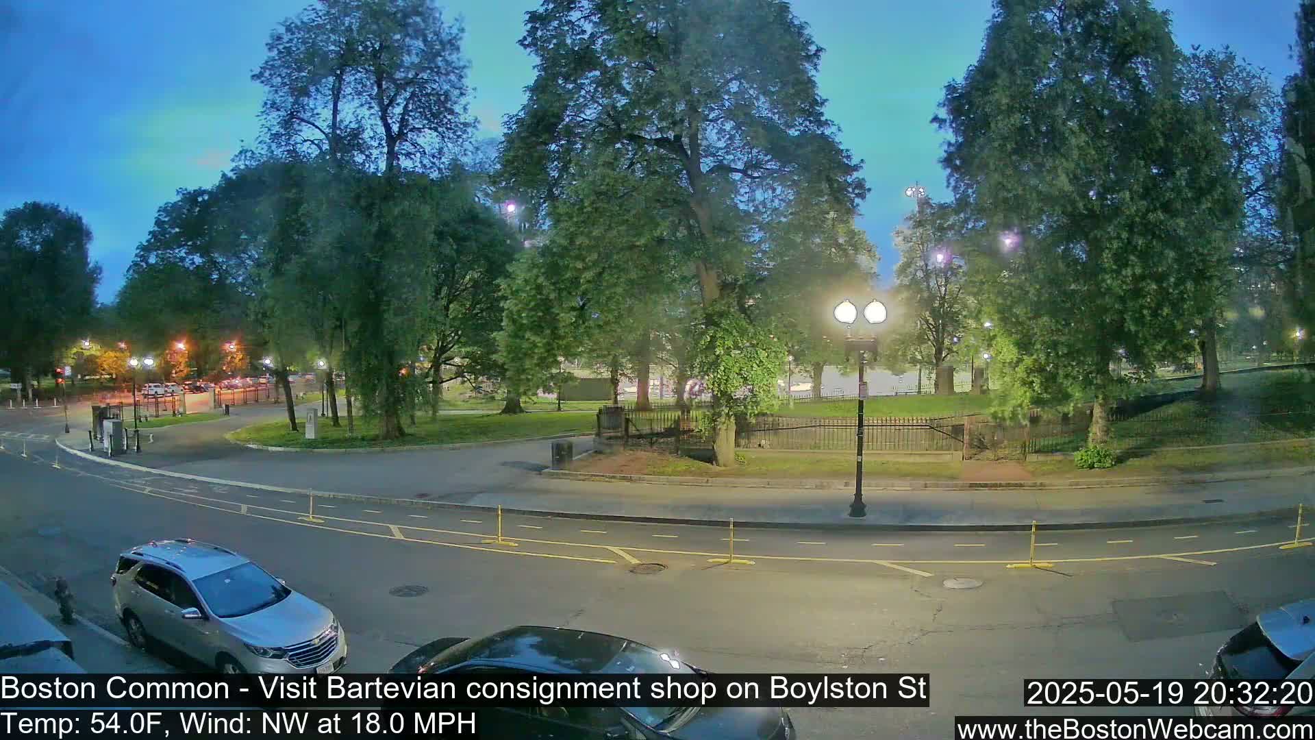 A street scene at dusk shows a park with trees and a roadway with cars parked along the curb under a mostly clear, twilight sky with a northwest wind.