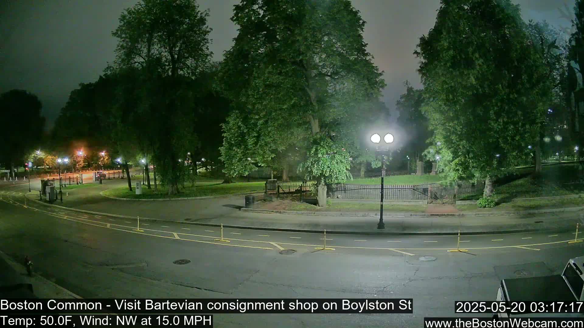 A nighttime view of a park, illuminated by streetlights, with a road curving through the scene under a cloudy sky, and a 15 mph northwest wind.