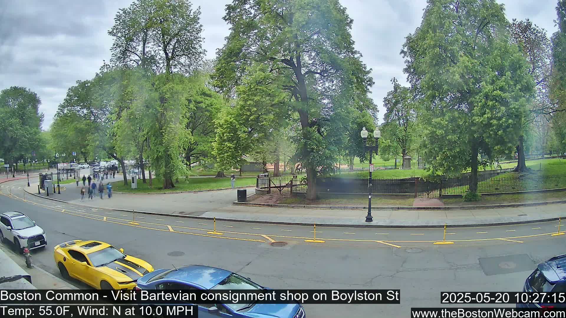 A partly cloudy day shows a street scene with a yellow sports car and other vehicles parked near a park entrance with people walking through a grassy area lined with trees.