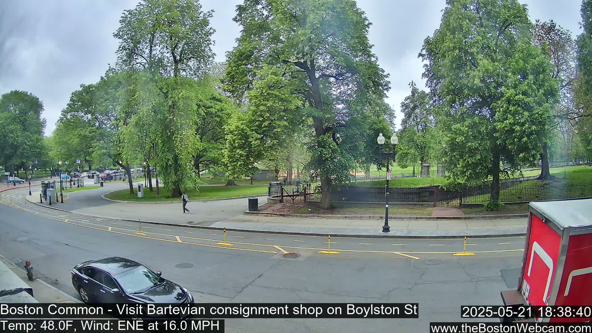 A partly cloudy day shows a street scene with parked cars, pedestrians, and lush green trees surrounding a park-like area, with a moderate wind.