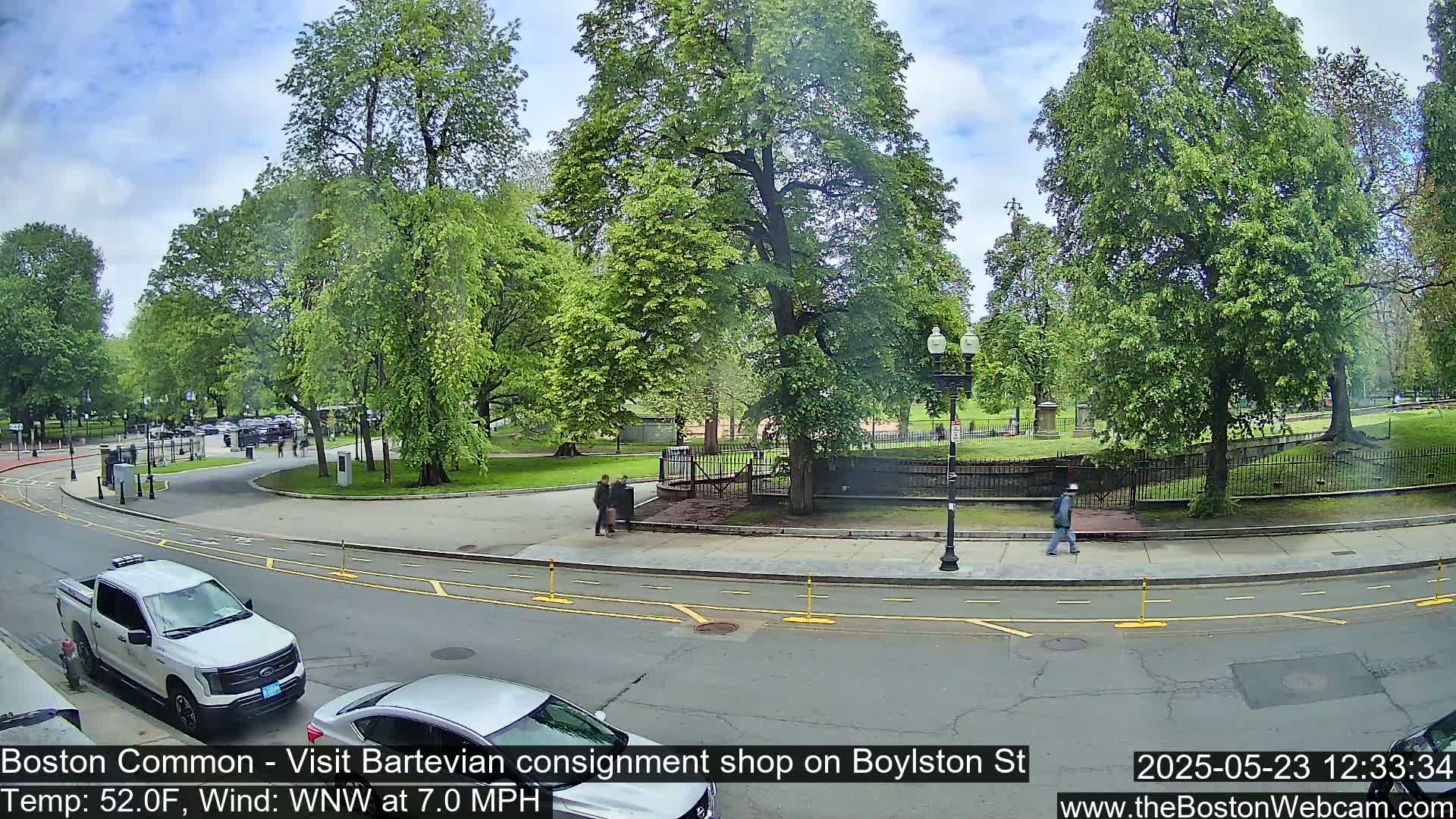 A partly sunny day shows a street scene with parked cars, pedestrians, and lush green trees lining a park.