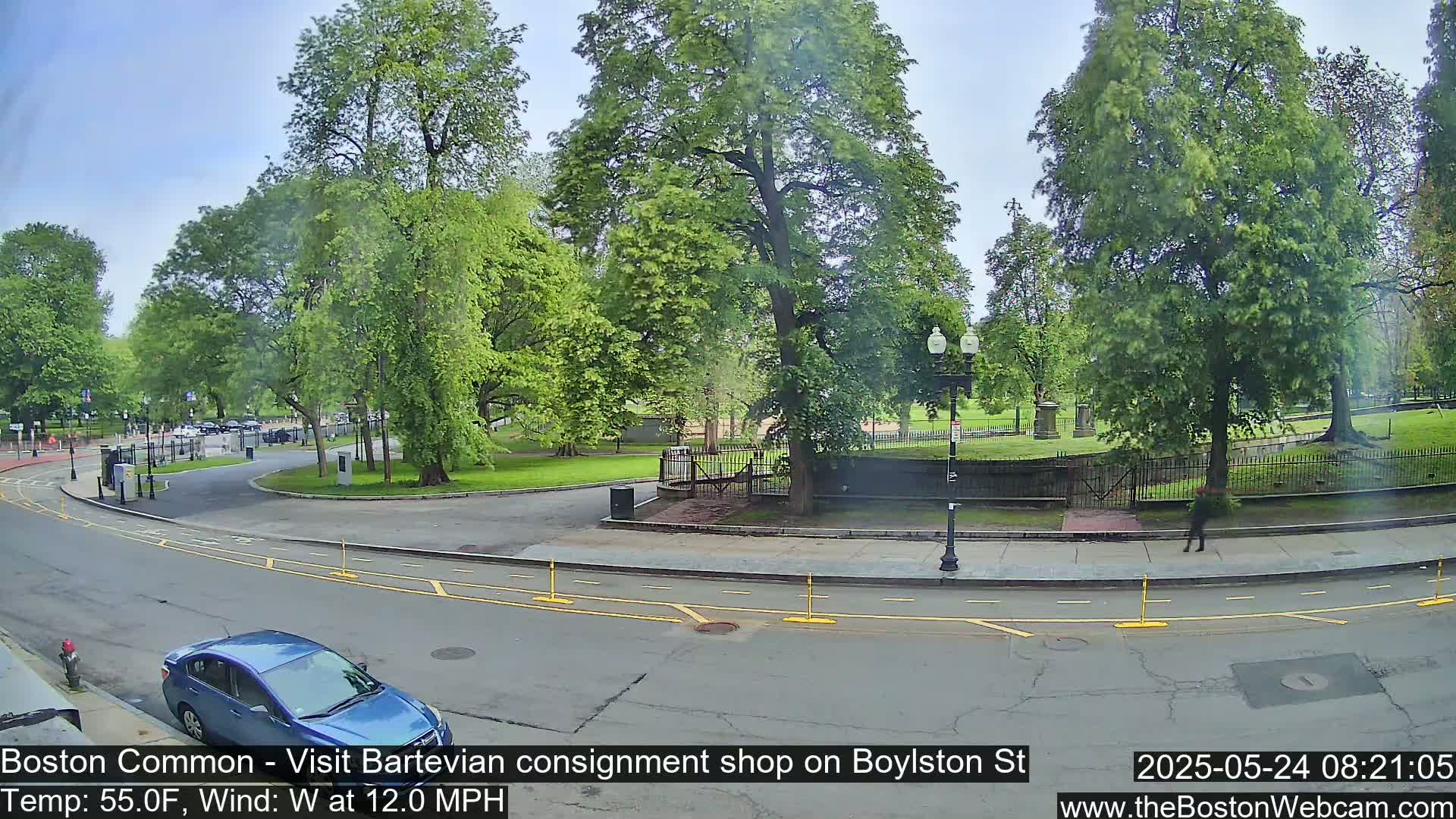 A partly sunny day shows a street curving around a park with lush green trees and a blue car parked near the curb.