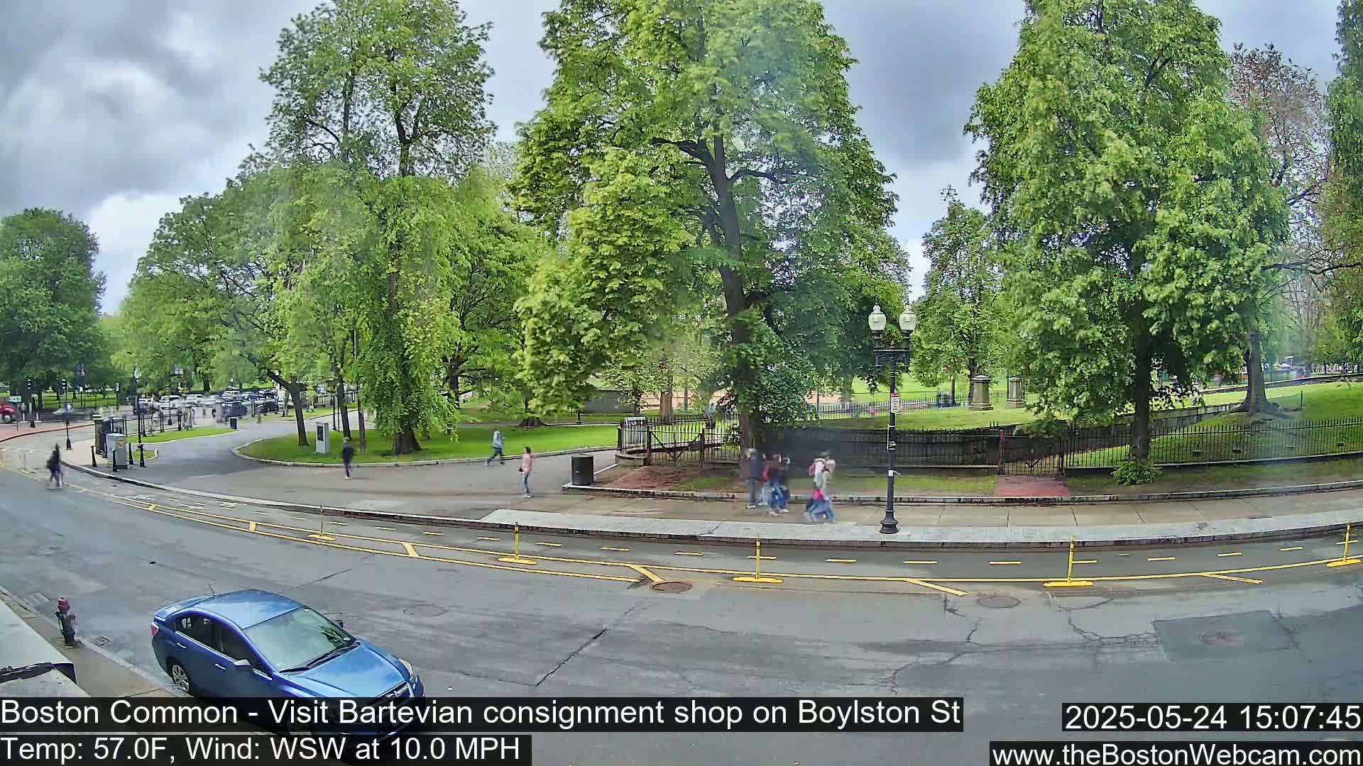 A partly cloudy day shows a street scene near a park with lush green trees, people walking, and a blue car parked on the side of a road.
