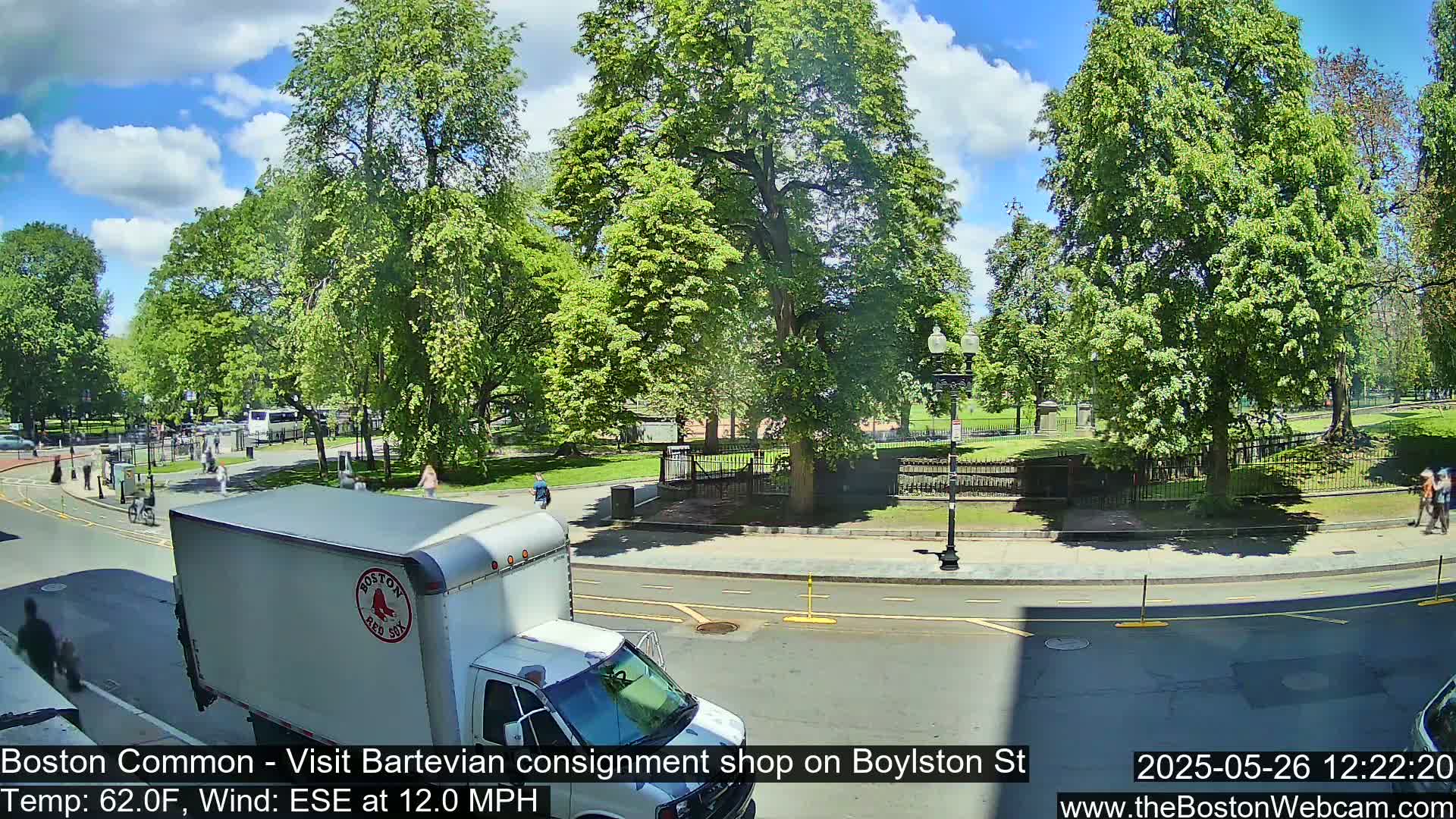 A sunny day shows a city street scene with a white van, pedestrians, and lush green trees bordering a park.