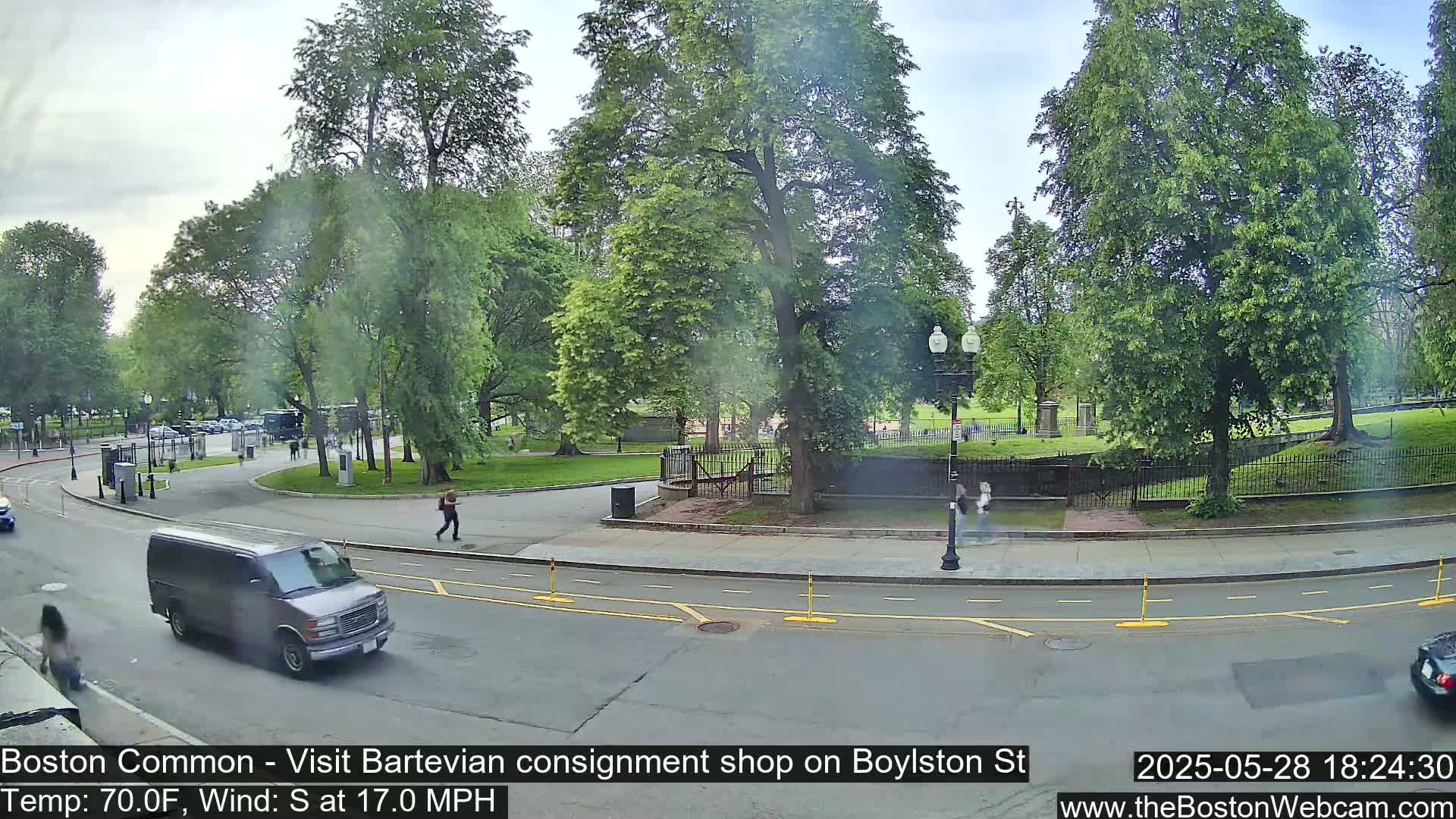 A city park with lush green trees and a paved road, under a partly cloudy sky, with a van and a few pedestrians visible.