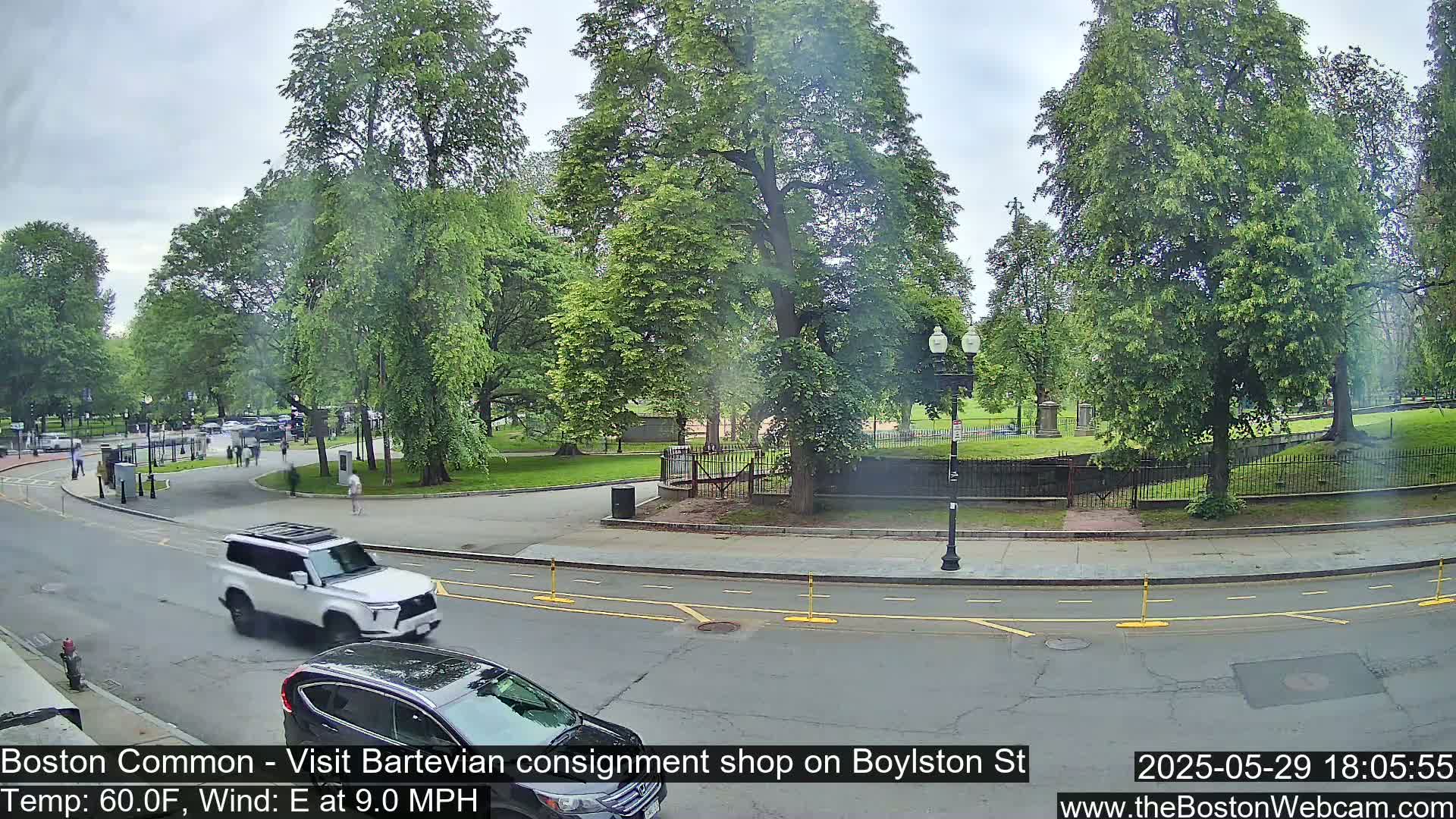 A partly cloudy day shows a street scene with cars driving past a park with lush green trees and a low stone wall.