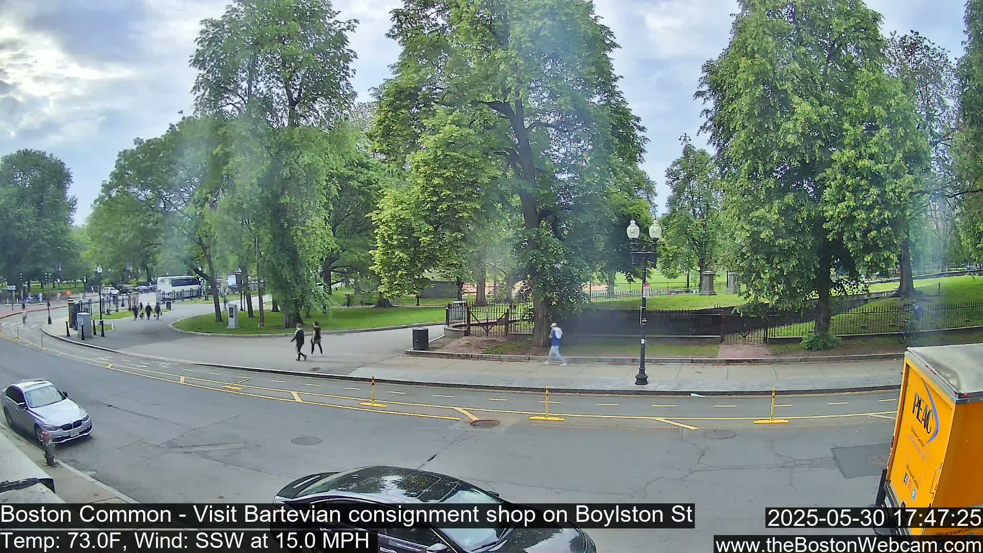 A partly cloudy day shows a street scene with pedestrians walking near a park filled with lush green trees.