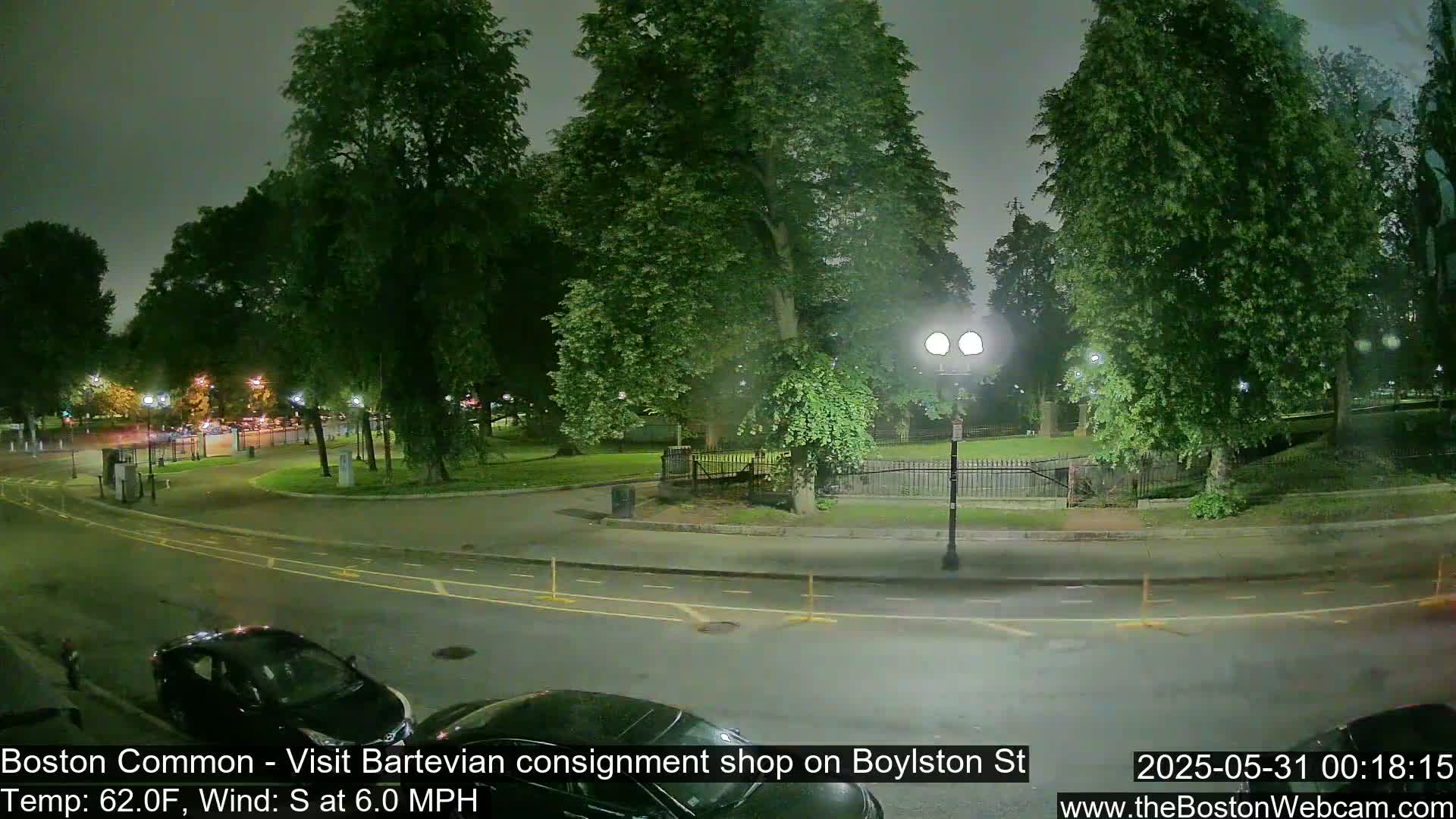 A nighttime view of a park and street, with cars parked along the curb and trees lining the roadway under a cloudy sky.