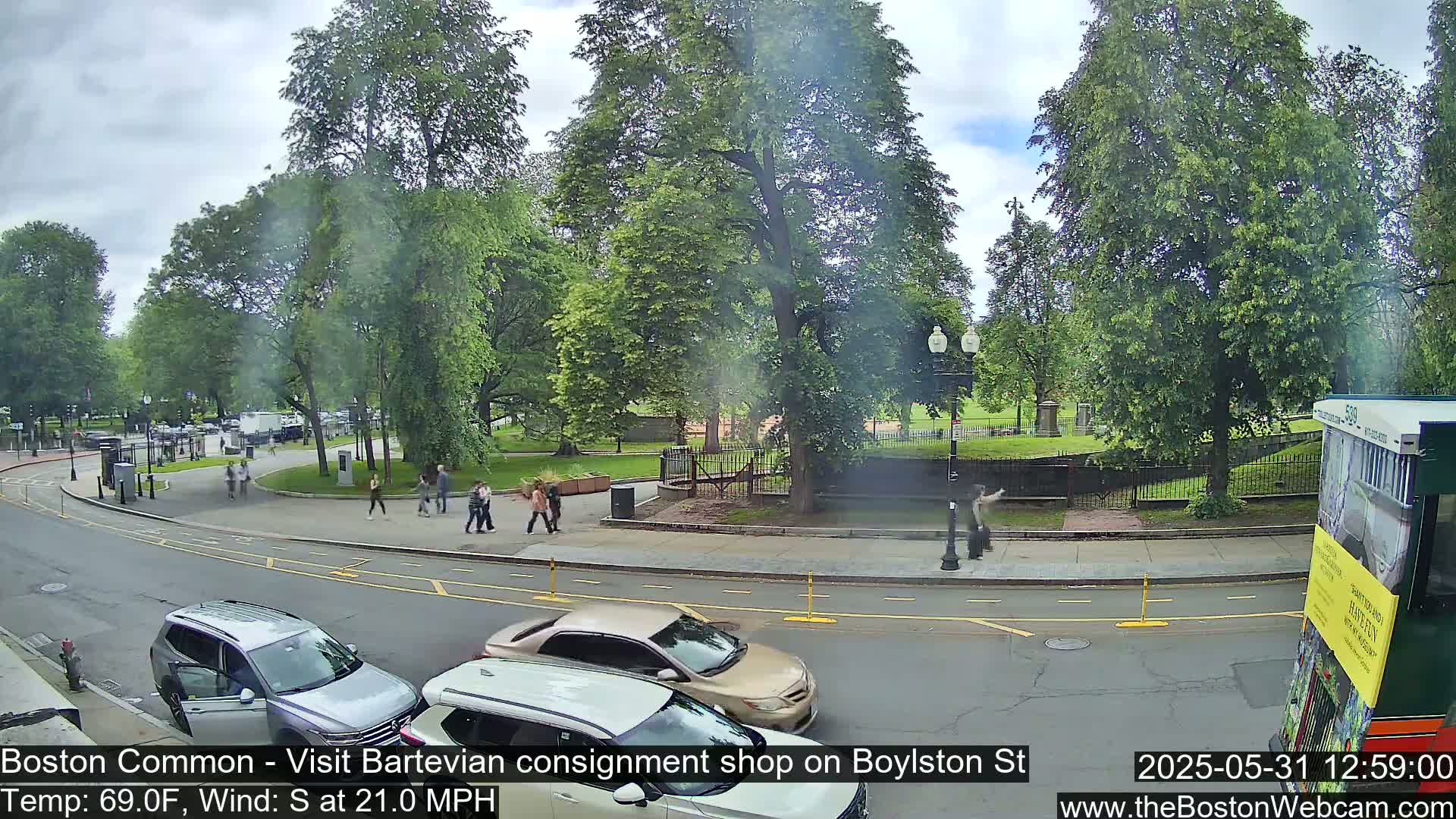 A partly cloudy day shows a city street scene with several pedestrians walking near a park filled with lush green trees and parked cars lining the road.