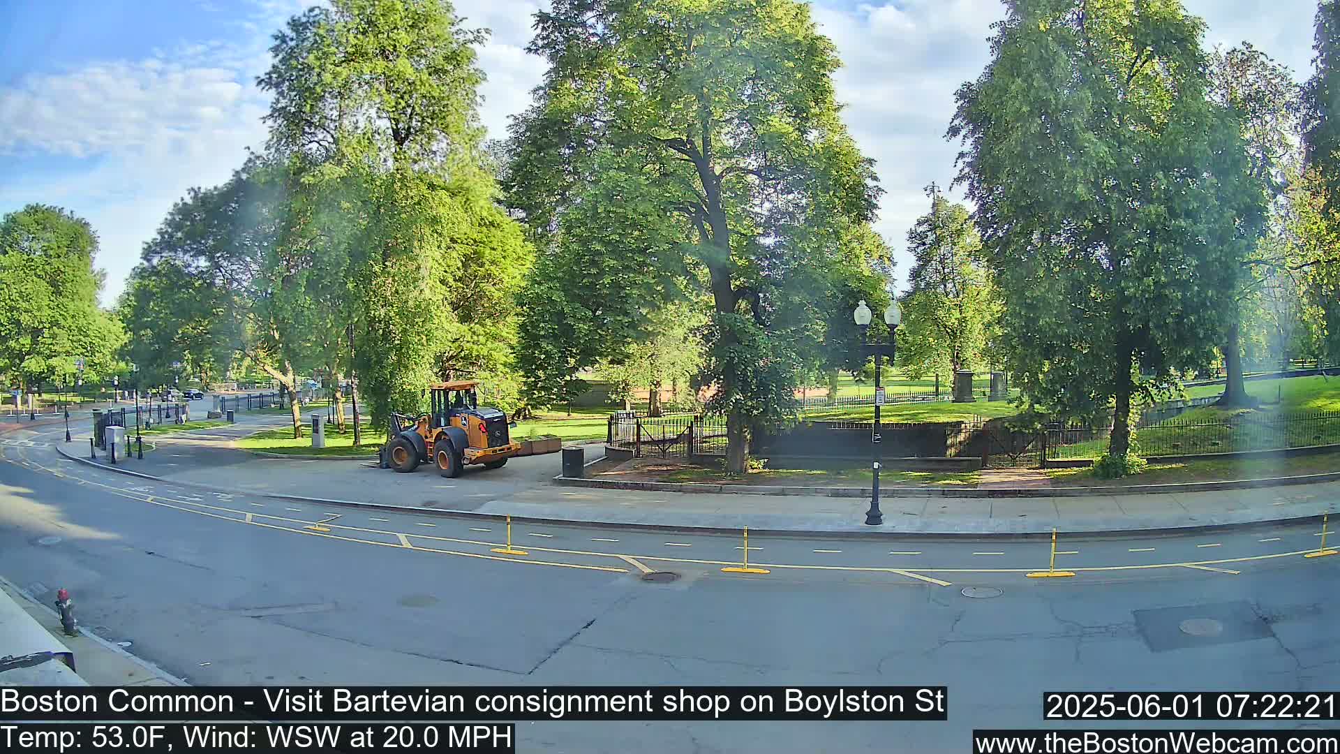 A partly sunny day with a 20 mph wind shows a yellow construction vehicle parked near a tree-lined road bordering a grassy park.