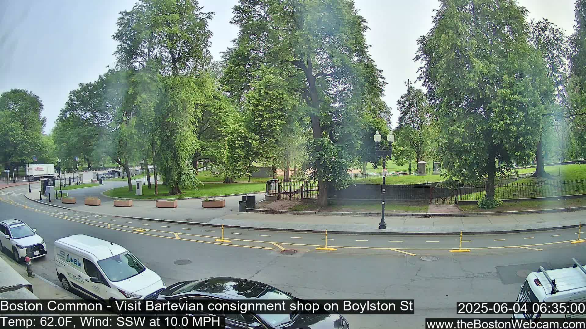 A partly sunny day shows a street scene with parked cars near a park with lush green trees and a low stone wall.