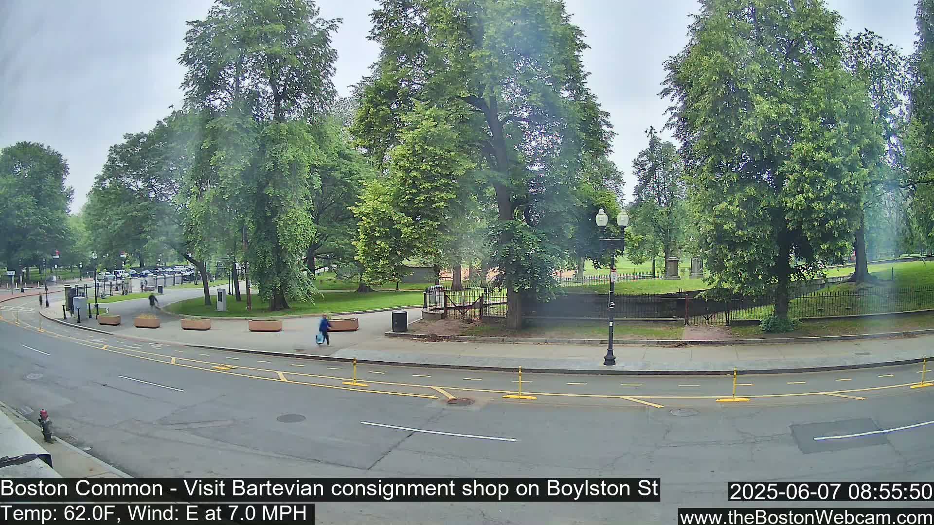A partly cloudy day shows a city street bordering a park with lush green trees and a few pedestrians.