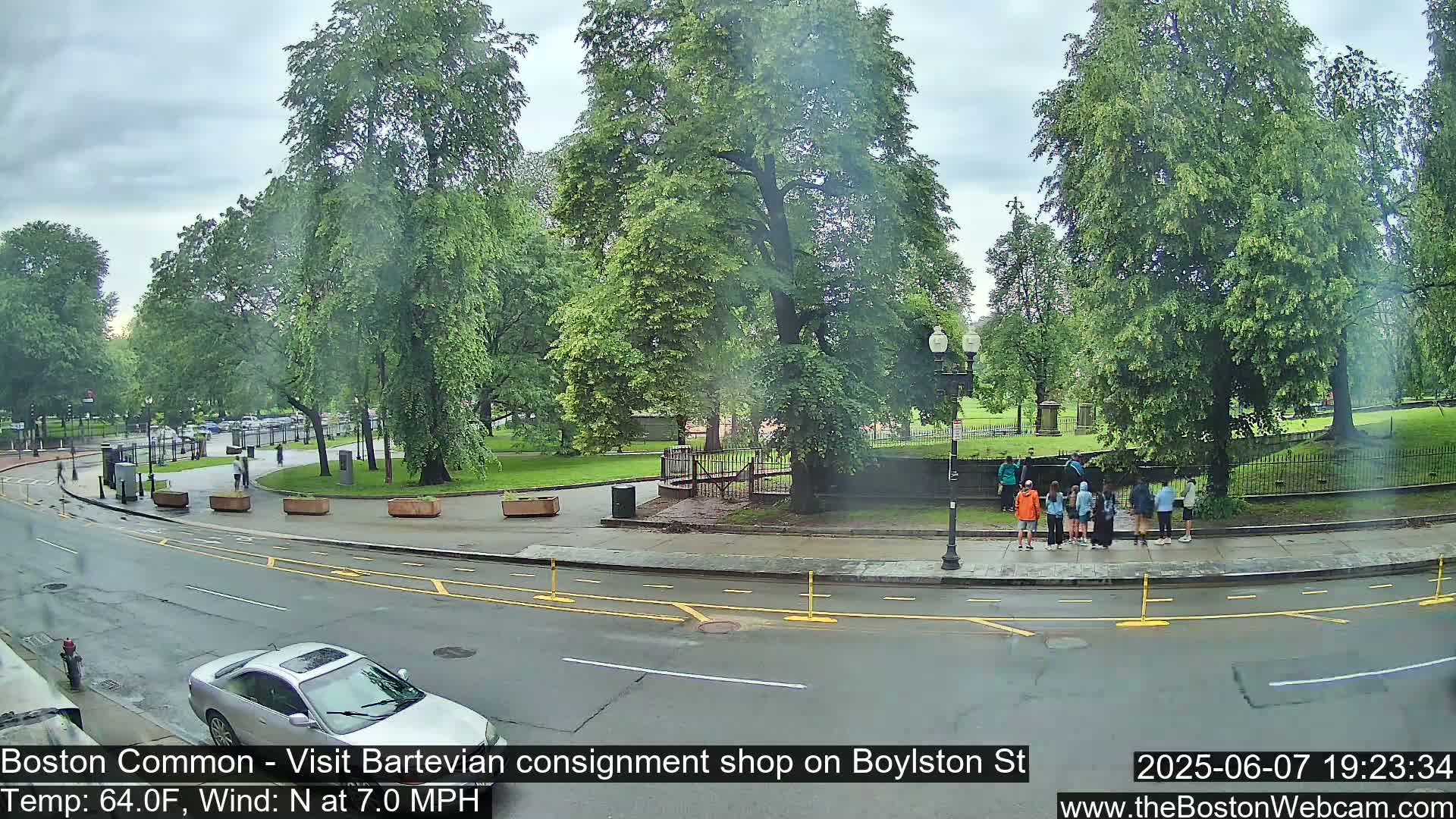 A wet street scene shows a park with lush green trees, several people standing near a fence, and a silver car parked near a roadway under an overcast sky.