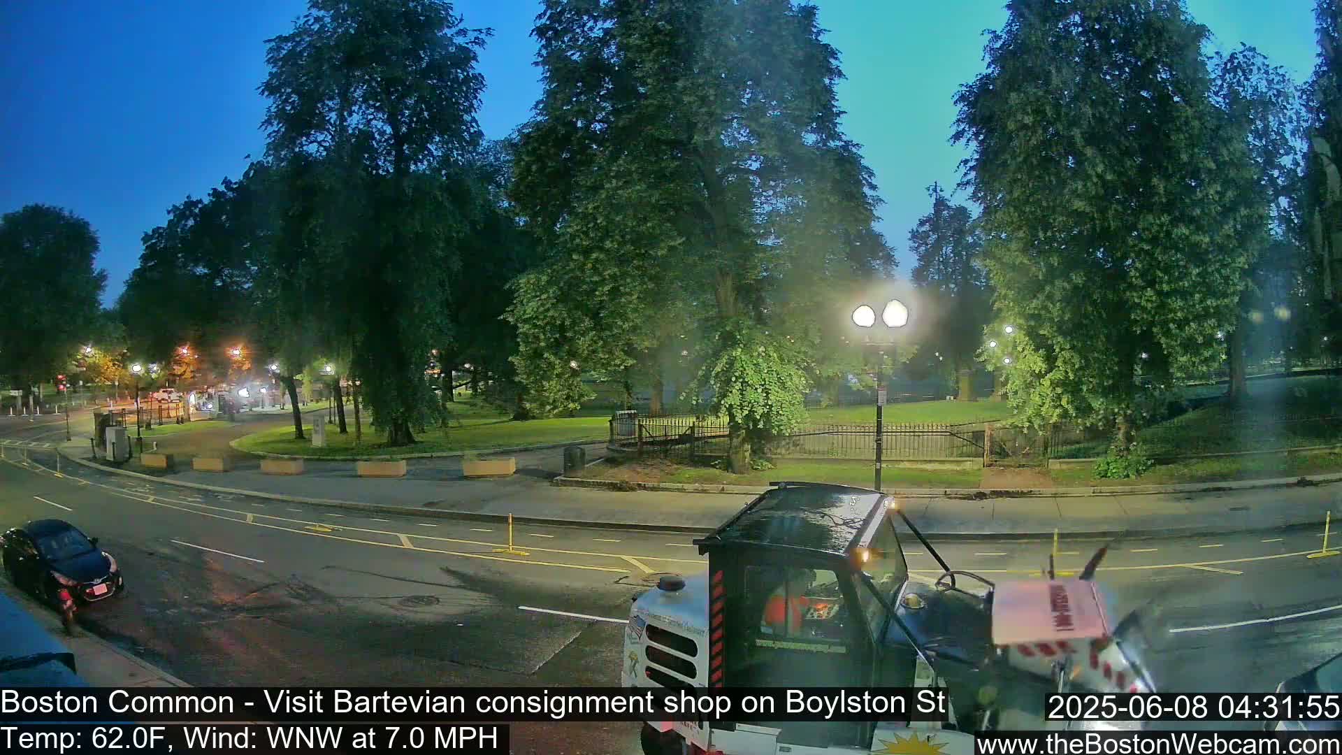 A street scene at dusk shows a park with trees and a streetlight, a vehicle in the foreground, and a car parked on the side of the road.