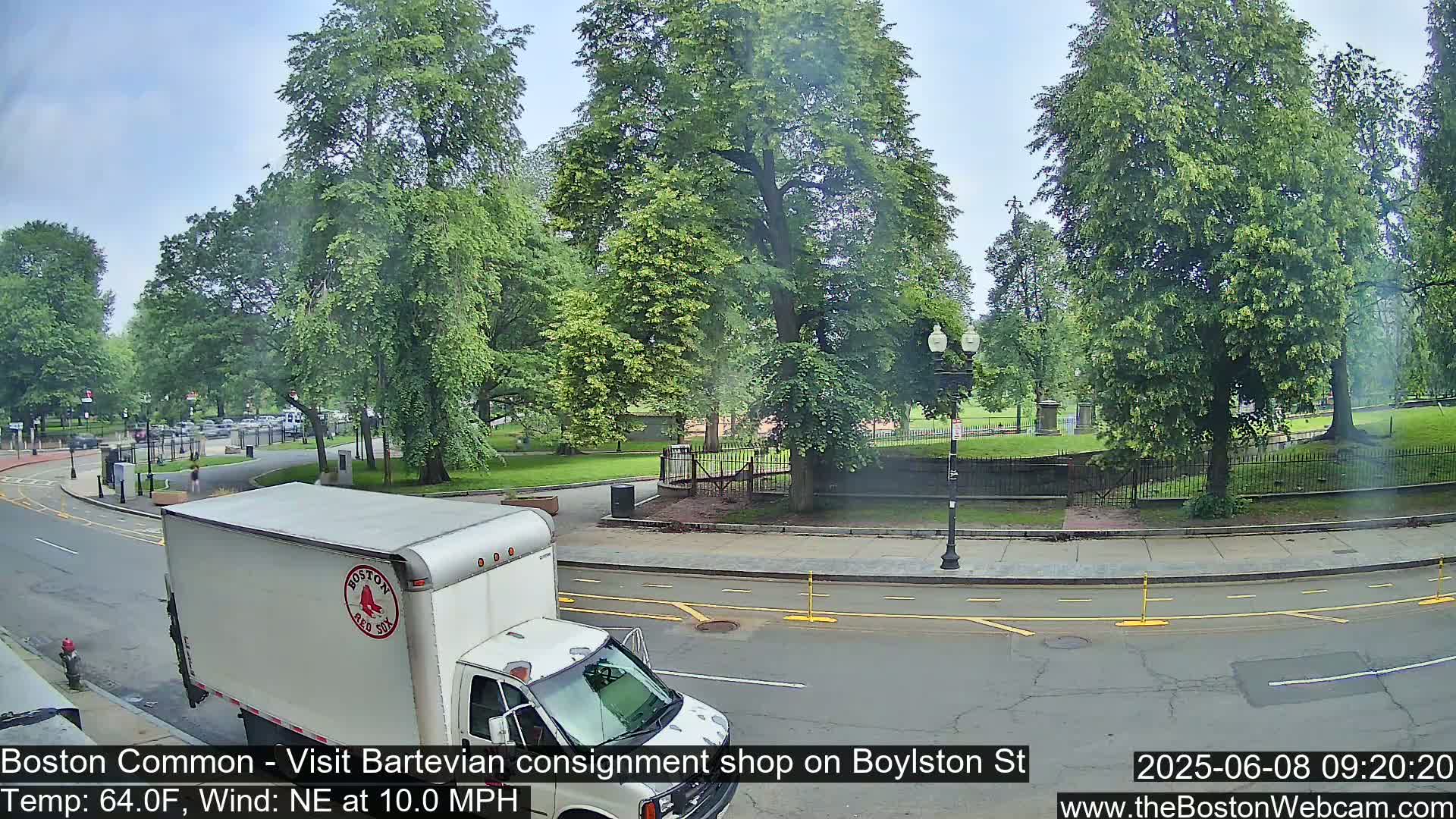 A white box truck drives past a park on a partly cloudy day with a northeasterly wind of 10 mph and a temperature of 64°F.